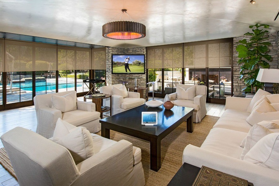 A living room with a Crestron touchscreen on the coffee table, partially drawn motorized shades, and golf on the TV.
