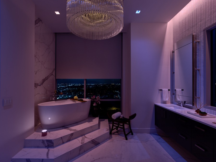 A bathroom illuminated in violet with a soaking tub and motorized shades lowered over the windows. 